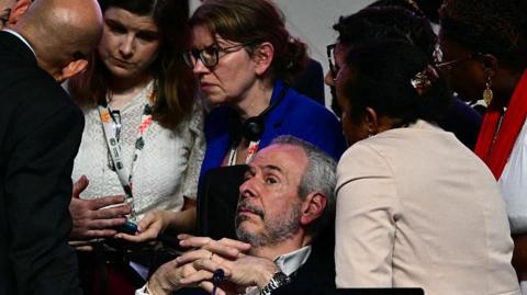 A bearded man, COP president Andre Correa do Lago lies back as several others surround him, looking worried, during  a critical moment in the COP30 talks