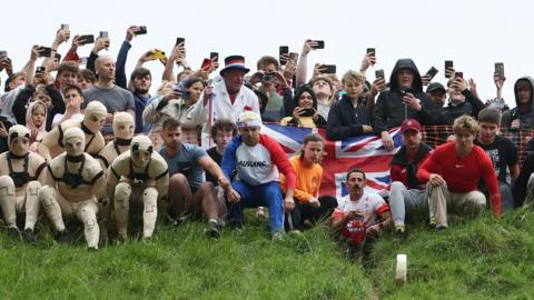 A crowd of people perch at the top of a steep hill as a wheel of cheese is released. YouTuber Zac Alsop's team of five stuntmen wear skin coloured catsuits to the left of the picture. YouTuber Tom Kopke wears a red Nike top and a khaki trousers. 