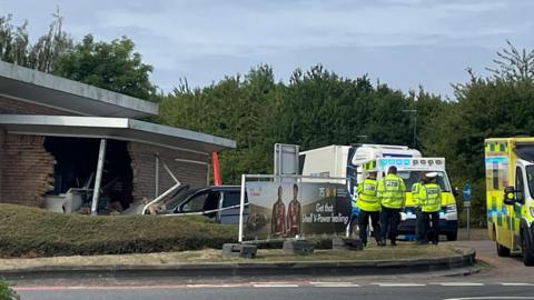 A damaged brick building with a damaged car beside it. The car has cashed into it. Four police officers in hi-vis jackets are standing on a grass verge to the side of the damaged building. There is an ambulance on the right of the picture.