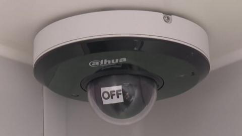 A ceiling-mounted internal CCTV camera. In front of its lens is an automatically movable sign that says "Off".