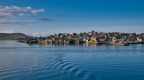 Houses and other buildings on the waterfront at Lerwick. There are ripples on the surface of the sea and the sky is blue.