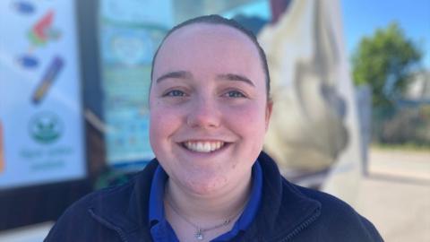 A woman with her dark hair pulled back and a nose ring faces the camera in an outdoor setting, wearing a blue top under a dark fleece. The background is softly blurred, showing signage, light-colored structures, and greenery in bright daylight.