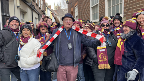 The picture shows more than a dozen men and women smiling at the camera. The majority are wearing burgundy bobble hats, and matching scarves. The man in the middle is wearing a red and white striped scarf around his neck, which he is holding out with his arms. They are standing in front of a row of houses.