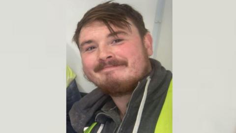 Dean Green is smiling at the camera. He has brown eyes, brown hair and a brown beard and moustache. He is wearing a high-viz jacket over a black hoodie.