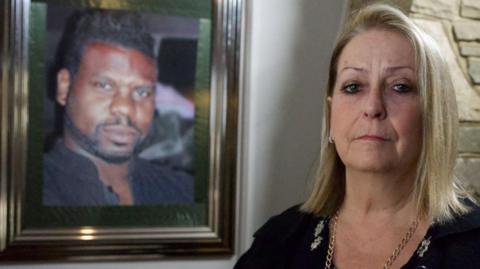 Carole Charles, a middle-aged woman looking sad. A photo of her husband is visible on the wall behind her. She is made up smartly and wears a black top and a gold-coloured chain. She has blonde hair coming down to her shoulders.