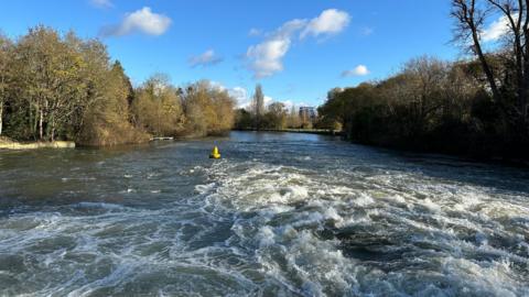 A river dominates the image with the water having been buffeted - possibly from a boat which is out of shot. It's a lovely sunny day with tree lines either side of the water.