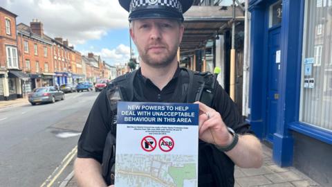 A uniformed police officer stands on a town-centre pavement holding a poster about new powers to tackle unacceptable behaviour in the area. The poster includes symbols related to alcohol and antisocial behaviour and a map of the affected zone. A row of brick-fronted shops and parked cars lines the street behind.