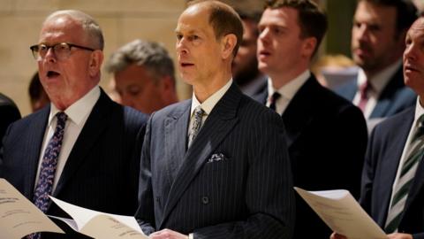 balding man in a pinstripe suit among a group of men singing a hymn