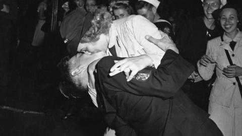 A woman wearing a naval cap, holding a cigarette as she kisses a uniformed sailor during the V-J Day celebrations in New York