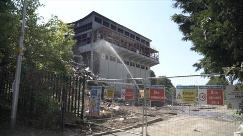 The former St John Fisher School in Chatham during demolition, with sections of the building removed, safety fencing in place and water being sprayed to reduce dust.