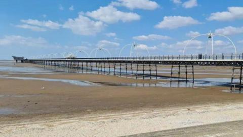 A generic photograph of the pier on Southport beach with blue sky and water pooled on the sand.