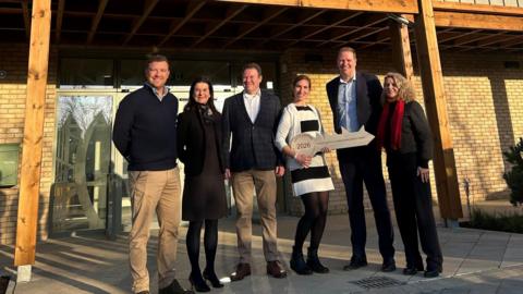 Three men and three women stand in front of a centre. They are all smiling and one is holding a large cardboard key. They are standing in front of a glass-panelled door under an awning. 