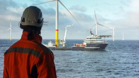 A person wearing orange overalls and a hard hat looks out over the North Sea. There is a ship next to a large wind pylon in the centre of the shot, with other pylons in the background. 