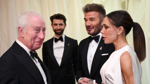 King Charles speaks with David and Victoria Beckham. Both men wear black tuxedos, while Victoria Beckham wears a sleeveless white gown and has her brown hair tied up in a ponytail.