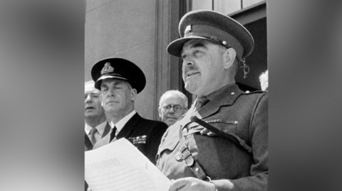Brig Snow is standing in full military uniform and looking away from the camera. He is holding a sheet of paper, and there are at least four men standing behind him. This is a black and white image.
