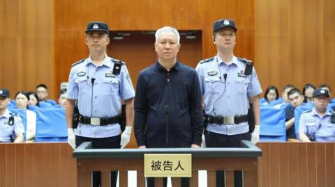 China Evergrande Group founder Hui Ka Yan takes the stand in a court in China. He has a blank look and is standing between two officers, with members of the audience in the background.
