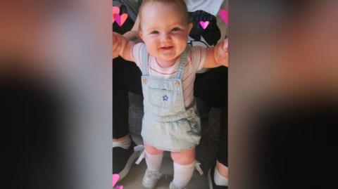 Ali Jayden is smiling at the camera, standing up and being held up by the hands of an adult wearing black trousers and black shoes with white on the bottom of the shoes. 
Ali has short gingery hair and eyebrows. She is wearing baby blue shoes and white socks pulled up to the knee. She is wearing a stripy pink and white tshirt and the photo has love hearts inserted around her head. 