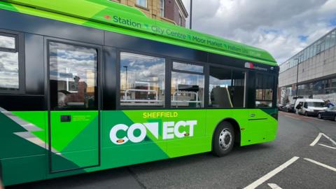 A large green electric shuttle bus, travelling through a city street