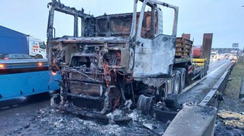 Burnt-out lorry on the hard shoulder.T he front is charred and the paintwork has gone.