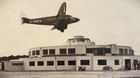 A sepia image of a circular building with a plane flying over the top of it.