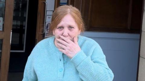 An older woman with frizzy red hair tied up in a ponytail and wearing a knitted blue cardigan walks out of a building. She is holding a hand over her mouth.