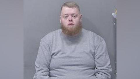 A custody image of Tyler McQuarrie, wearing a grey top and sitting looking straight at the camera. He has a ginger coloured beard and short fair hair.