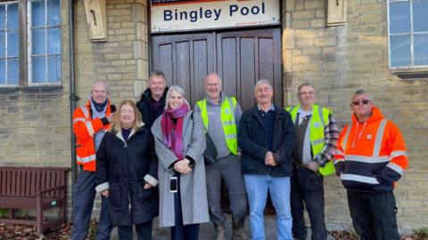 A line up of smiling men and women outside the wooden door entrance to Bingley Pool