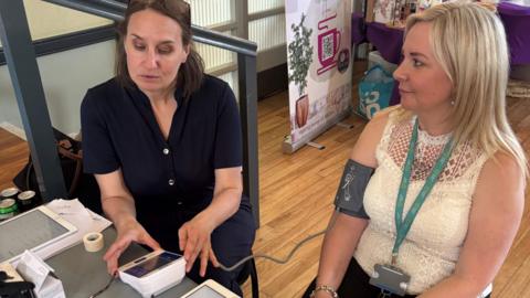 Dr Karen Forshaw is wearing a blue top and has long brown hair. She is taking the blood pressure of Sarah Chappell who has long blonde hair and is wearing a light top.