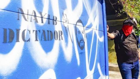 A student stands next to a graffiti against the government of Nayib Bukele in San Salvador