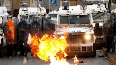 PSNI officers in riot gear and Land Rovers as a fire burns on the Springfield Road, Belfast 8 April 2021