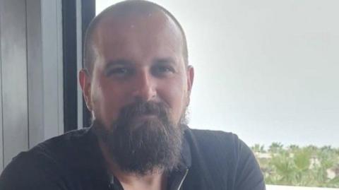 A bald man with a beard and dark shirt smiles as he stands in front of a window.