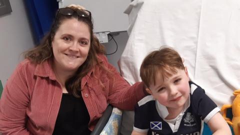 Rebekah, a woman with brown hair, wearing a pink shirt, sat with her young son Hector who looks up at the camera in a hospital room.