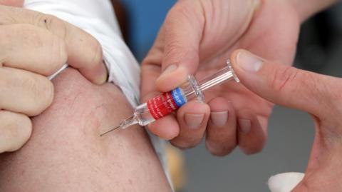 A medical professional injects a patient with a vaccine. The patient is seen lifting up their sleep while the injection is given.