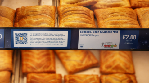 A range of baked savoury products can be seen on display in a Greggs store, with signs including 'freshly baked', 'adults need around 2000kcal a day', 'vegan festive bake' and 'cheese and onion bake'.