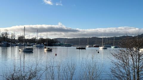 A general view of boats on the water at Ferry Nab in Windermere. The small sailing boats are stationary on the lake. In the background there are fells on the opposite side of the lake. The sky is blue with a few white clouds.