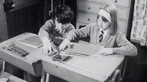 Black-and-white image of a boy and a girl sat at old-fashioned school desks in front of a chest of drawers
