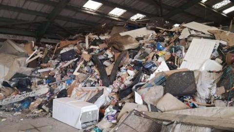 A large pile of household and construction waste, including mattresses, doors, furniture, appliances, timber and black bin bags, fills an industrial warehouse almost to the roof.