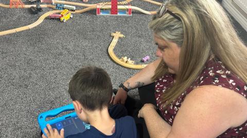 A woman sits behind a young boy who is playing on a tablet with a blue case. They are both looking at the screen. The back of his head is visible, he has short brown hair. A wooden train set is in the background of the image.