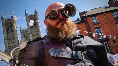 The man wears goggles, one bigger than the other, and has a big ginger beard.  He appears strapped up in some kind of costume, and stands on a sunny day in front of Lincoln Cathedral.