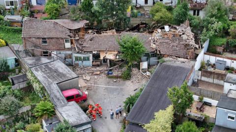 A partially collapsed building on Ryde Road, in the Elm Grove area of Brighton, has been cordoned off by emergency services. Firemen in bright orange uniforms gather alongside three casually dressed individuals to the south of the property. The building appears to have fallen from the centre of the roof, while the first floor remains intact. Dust and debris scatter the driveway and surrounding area. A triangular formation of houses, with their back gardens facing toward the partially collapsed building, envelop the site.