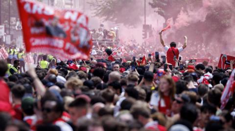 A dense crowd of Arsenal football supporters celebrates in a city street filled with red smoke. In the foreground, a large red banner is blurred, while in the mid-ground, a fan stands above the crowd with arms raised, holding a smoking flare.