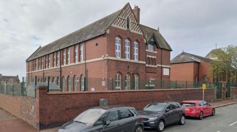 A general view of Sacred Heart Catholic Primary School seen from the street. The two-storey building has large windows on both floors and is built of red brick, with a chequered red and cream motif at the top. A wall topped by a green metal fence surrounds the building.