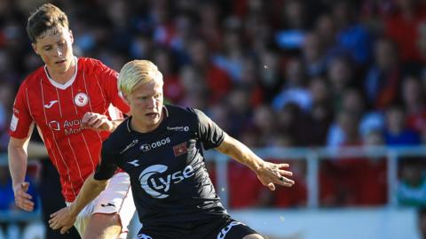 Harald Nilsen Tangen is pictured in a black playing shirt, holding off an opposition player who wears red. The background is blurred, but it is full of fans. 