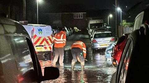 Picture shows flooding in a residential street. Welsh water engineers stand in front of a Welsh Water van working to sort the problem. 