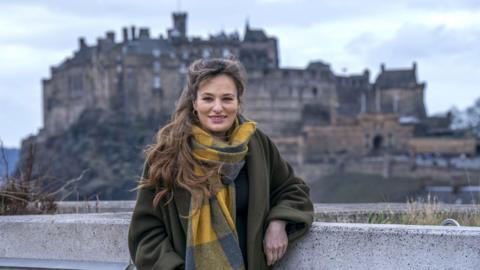 Nicola Beneditti standing and smiling with Edinburgh Castle far in the background. She is wearing a green coat with a large yellow and blue scarf, and is leaning against a wall.