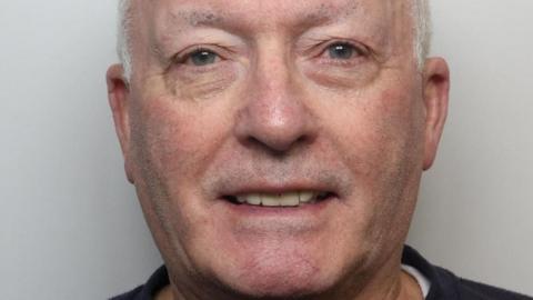 The image is a close-up portrait of an older individual with a bald head. He has blue eyes and is giving a slight smile. He is wearing a dark-coloured shirt with a round neckline. The background is plain and light in colour.