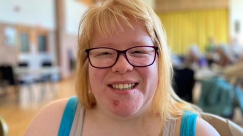 A woman with shoulder-length blond hair and purple glasses. She is wearing a light blue dress and is smiling. She is sitting down in a community hall with people behind her. The background is out of focus.