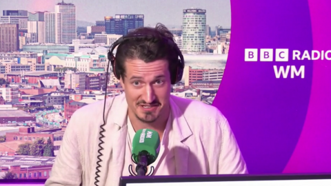 A man with dark brown hair, wearing a white t-shirt and jacket, sits at a desk with black headphones on. He is leaning towards a green microphone which says 'BBC Radio WM' on it. The background shows a picture of Birmingham on the left and 'BBC Radio WM' on a purple background on the right.