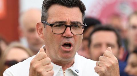 Andy Burnham is captured mid-gesture with both hands raised to chest level, his fingers curled into fists. He is wearing dark-framed glasses and a white zipped top. His mouth is open in mid speech.