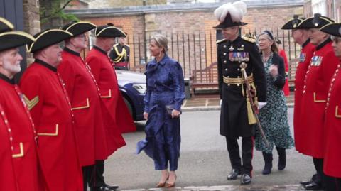 Chelsea Pensioners in their distinctive red tunics stand in two rows as an honour guard as the Duchess of Edinburgh walks into the Royal Hospital, accompanied by a man in formal uniform with rows of medals on his chest. He is wearing a plumed hat.